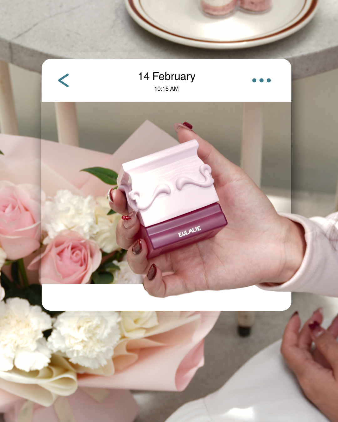 A hand holding a pink and plum fragrance box in front of a bouquet, inside a '14 February' message card (slide 2 of 4)